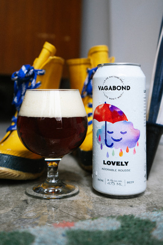 Canette et verre de bière Vagabond Lovely – Adorable Rousse 4,1%, bière ambrée artisanale servie sur un plancher mouillé avec bottes de pluie jaunes en arrière-plan, ambiance cozy et pluvieuse.