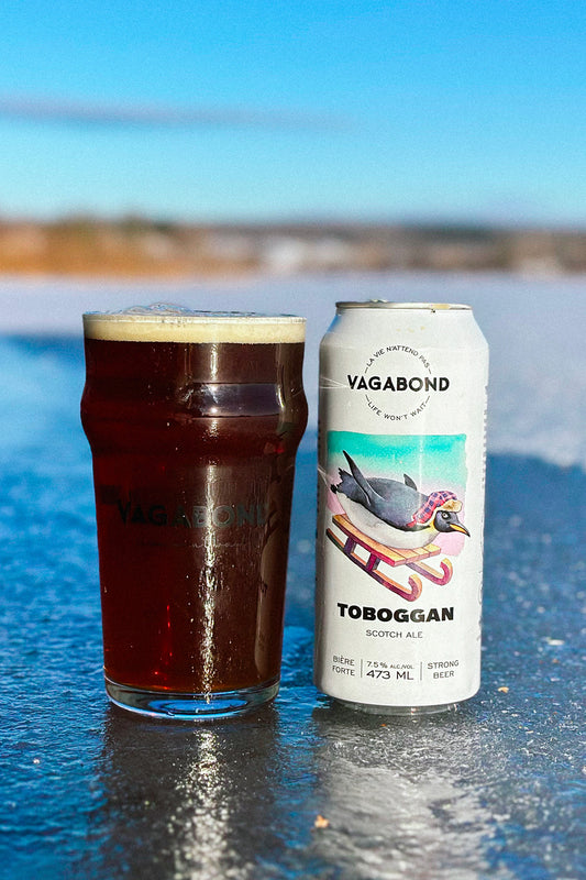 Canette et verre de bière Vagabond Toboggan – Scotch Ale 7,5% dégustée sur un lac glacé en hiver, bière forte brune artisanale riche et réconfortante.