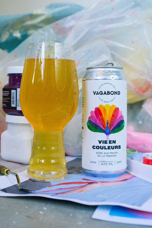 Canette et verre de bière Vagabond Vie en Couleurs – sûre aux fruits de la passion 4,5%, posés sur une table d’artiste avec pinceaux et peinture, ambiance créative et colorée.