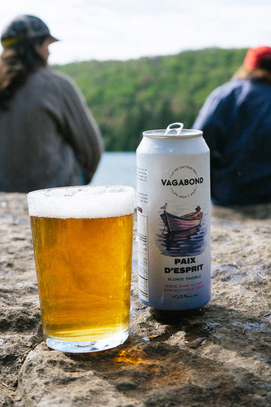 Canette de bière sans alcool Vagabond Paix d’Esprit et verre de bière blonde posés sur une roche, avec des personnes assises au bord d’un lac en arrière-plan.