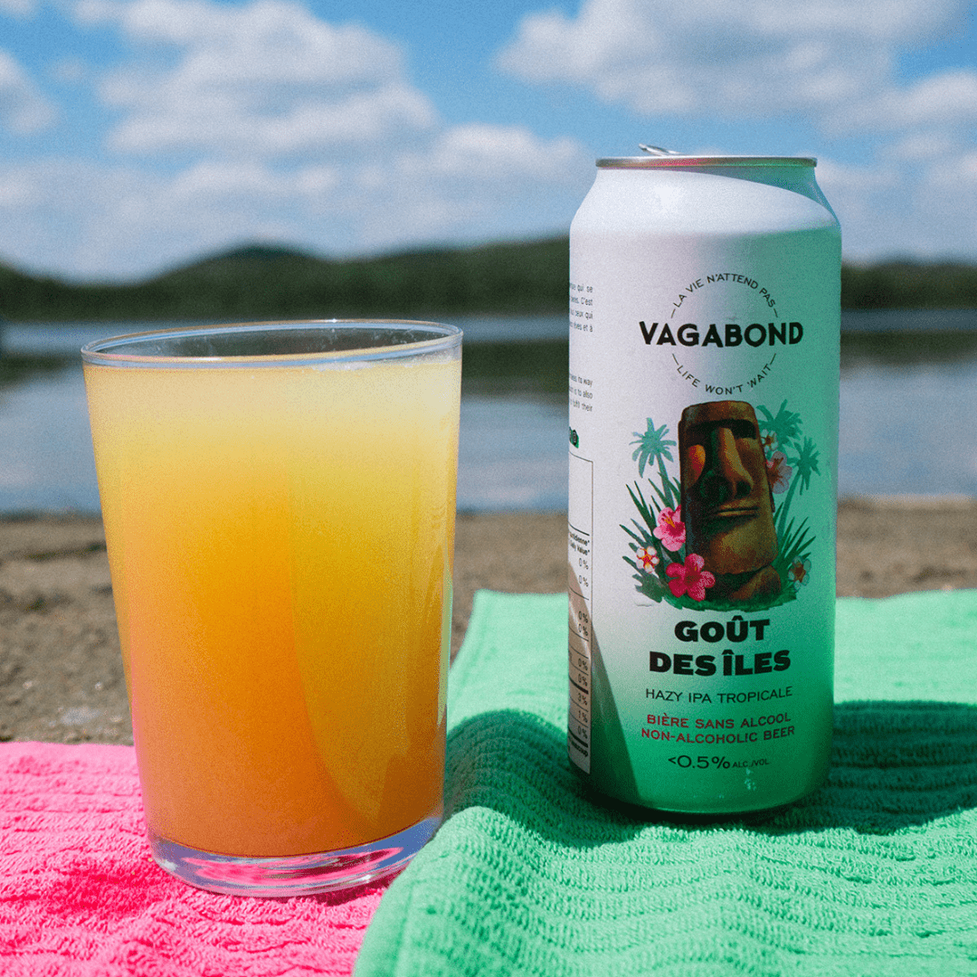 Canette de bière sans alcool Vagabond Goût des Îles avec un verre de bière sur une plage avec des serviettes de plage.