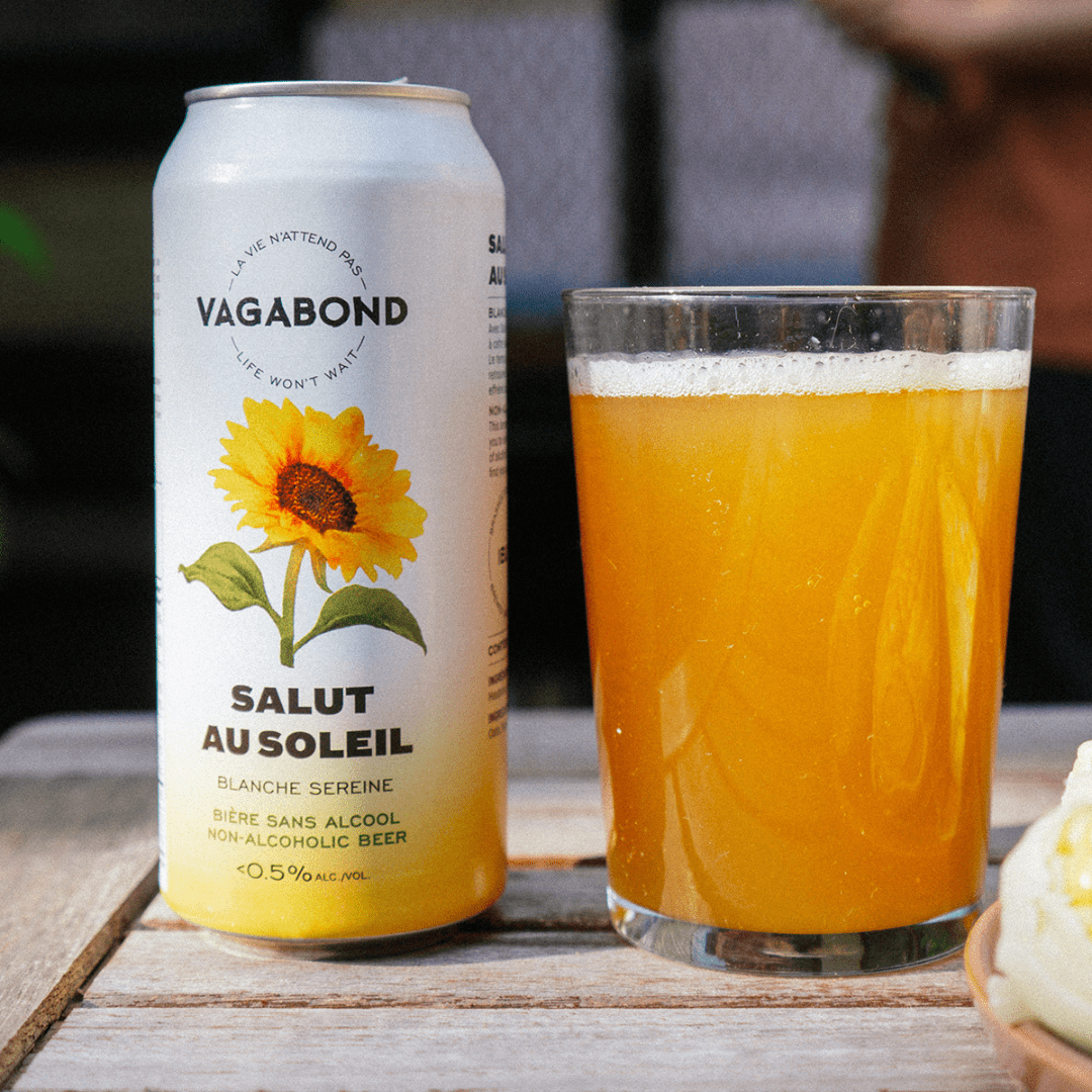 Canette de bière sans alcool Vagabond Salut au Soleil avec un verre de bière et une personne au BBQ en arrière-plan.