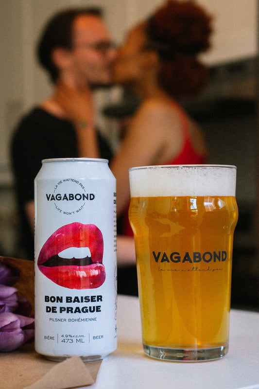 Canette et verre de bière Vagabond Bon Baiser de Prague – Pilsner bohémienne dorée 4.9%, photo lifestyle avec un couple qui s’embrasse à l’arrière-plan.