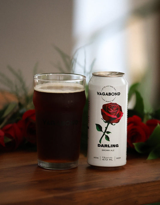Canette et verre de bière brune Vagabond Darling – Brown Ale 5% douce et maltée, posés sur une table avec roses rouges en arrière-plan