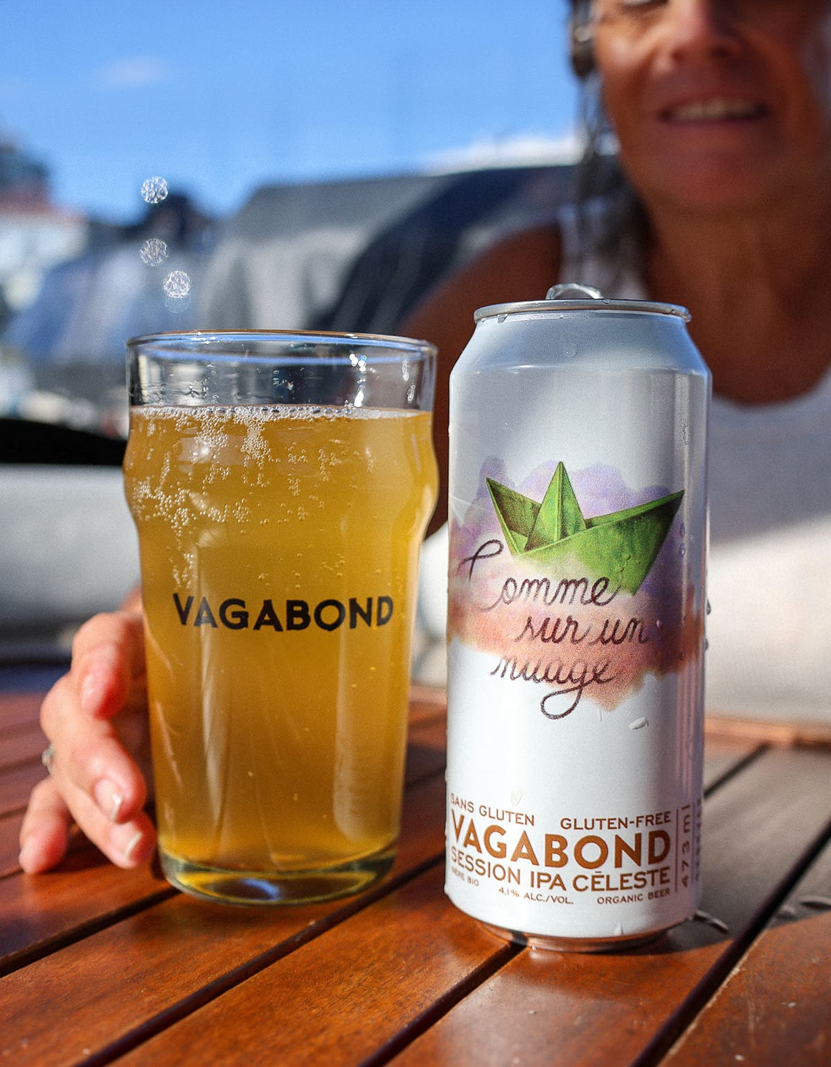 Verre et canette de bière sans gluten Vagabond Comme sur un Nuage – IPA légère et bio dégustée sur une terrasse ensoleillée avec une dame qui sourit
