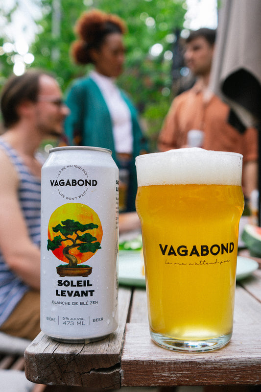 Bière Vagabond Soleil Levant – Blanche de blé zen servie sur une terrasse entre amis, avec la canette illustrée d’un bonsaï devant un soleil levant et un verre doré mousseux, ambiance estivale et conviviale.