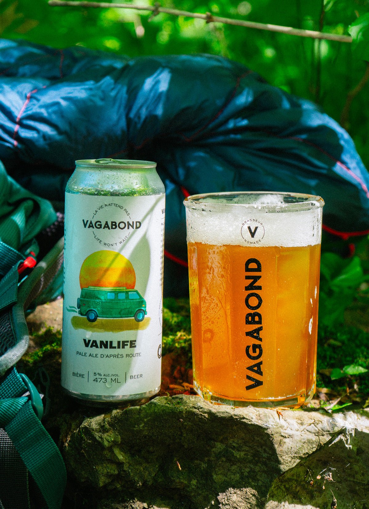 Canette et verre de bière Vagabond Vanlife – Pale Ale d’après route, en forêt sur une pierre, avec sac de couchage et sac à dos, ambiance d’aventure et de plein air.