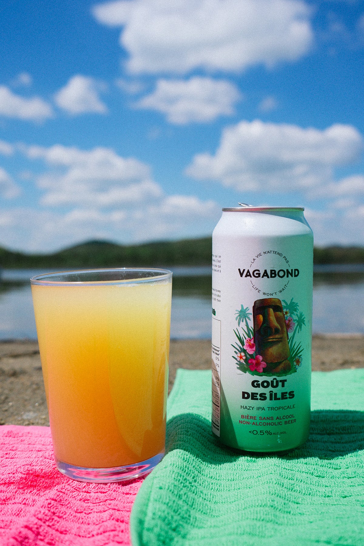 Canette de bière sans alcool Vagabond Goût des Îles et verre de bière tropicale posés sur une serviette au bord d'un lac.